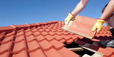 Construction worker tile roofing repair Roof repair, worker with yellow gloves replacing red tiles or shingles on house with blue sky as background and copy space.